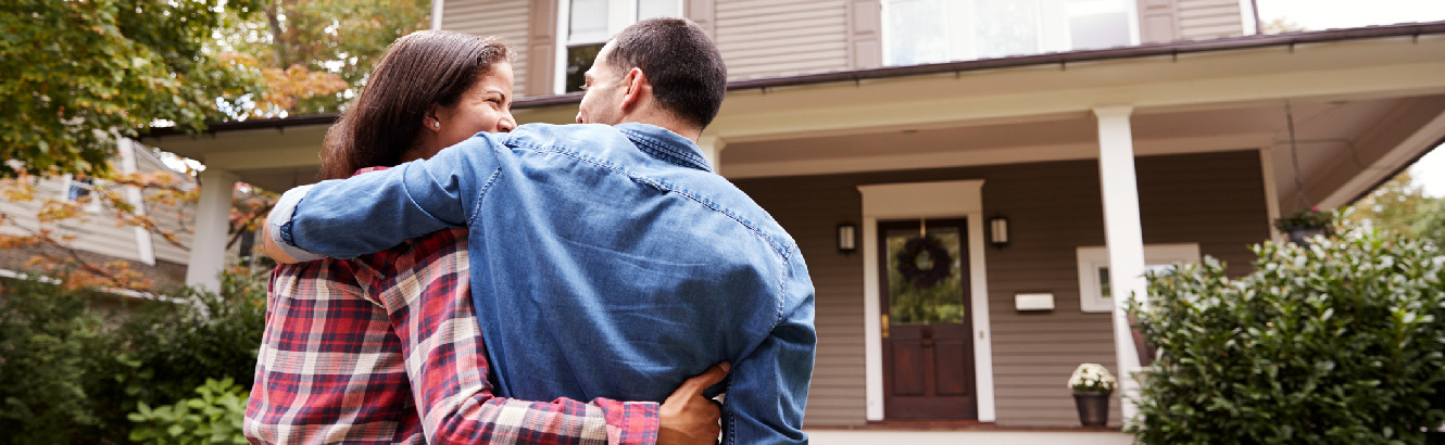 A husband and wife are embracing outside in front of a well-manicured lawn and nice house. Their backs are to the camera. 